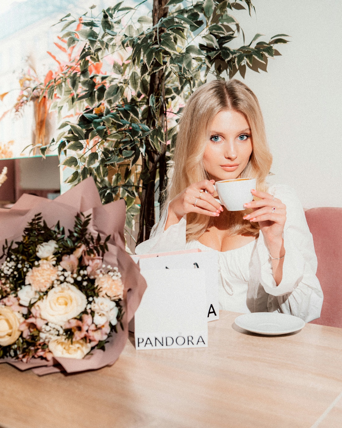 Blonde woman sips tea beside flowers and a pandora bag.