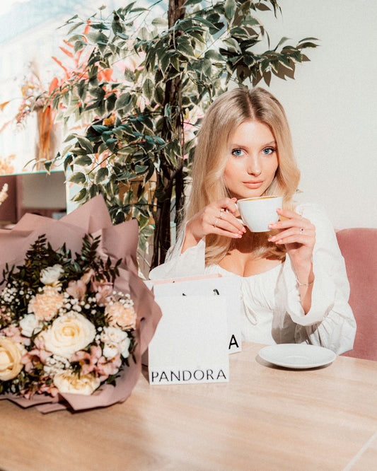 Blonde woman sips tea beside flowers and a pandora bag.